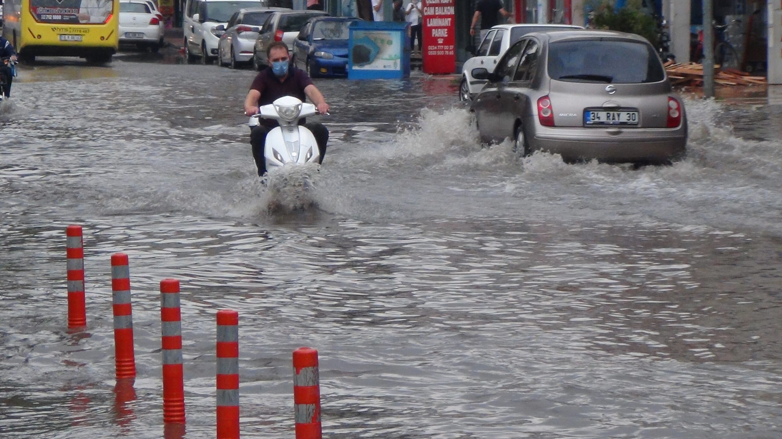 Su basmış bir sokakta yolun sağ tarafında araçlar dururken, sokağın ortasında beyaz motosikletli adam aracını sürüyor