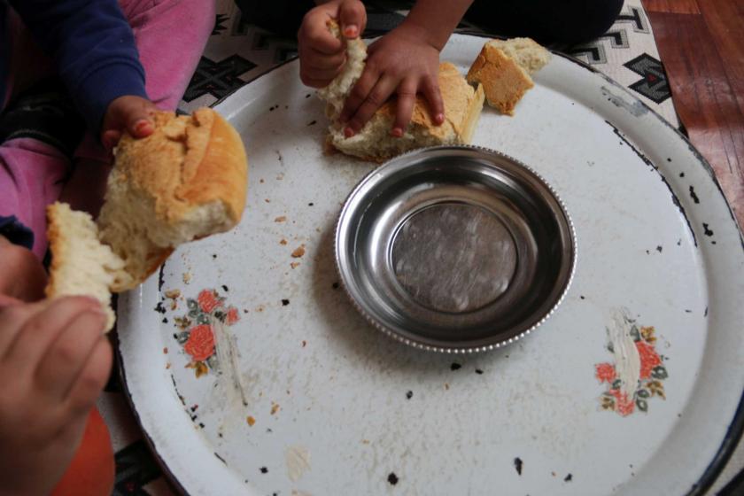 Boş bir sini içersinde boş bir metal tabak ve sini üzerine uzanmış iki çocuk eli ekmek parçalıyor