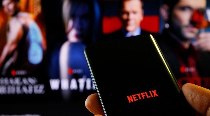 Netflix anasayfasının açık olduğu telefonu tutan bir kişi, arka zeminde de Netflix'e ait içeriklerin flu görüntüsü yer alıyor