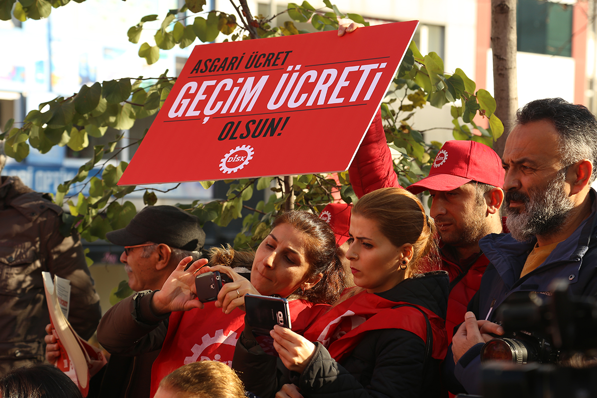 Kırmızı zemin üzerine yazılmış, 'Asgari ücret geçim ücreti olsun' dövizi taşıyan kırmızı şapkalı bir erkek ve hemen önünde iki kadın telefonlarıyla fotoğraf çekiyor