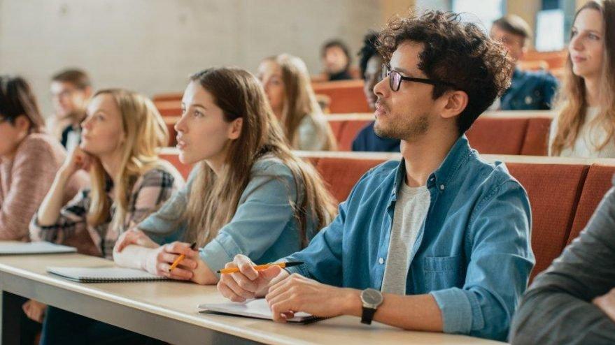 Üniversite amfisinde kadın ve erkek öğrenciler dikkatli bir şekilde ders dinliyor. Bazı öğrencilerin elinde not defteri ve kalem var.