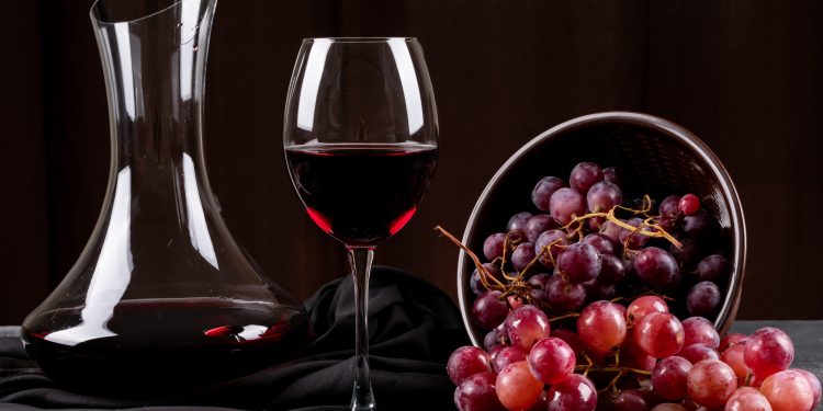 Bir şişe içinde duran şarap ve bardağın yarısına kadar dolu kırmızı şarap bulunuyor. Hemen yanı başlarında ise bir sepet içinde kırmızı üzüm bulunmakta.