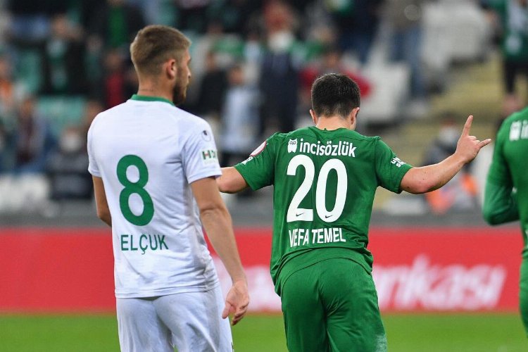 Yeşil beyaz formalı iki futbolcu sahada görülüyor. Biri 8 numaralı oyuncu Selçuk ve 20 numaralı Vefa Temel.