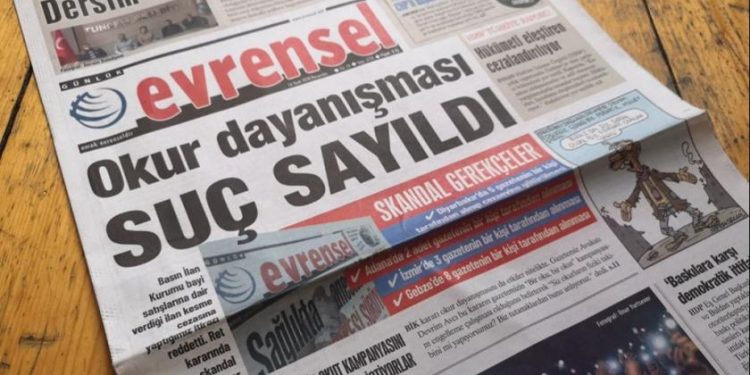 Tahta masanın üstünde Evrensel gazetesi açık duruyor. Gazetenin manşetinde "Okur dayanışması suç sayıldı" haberi yer alıyor