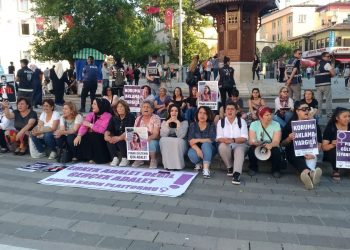 Çok sayıda kadının önünde bir pankart bulunuyor. Kadınlar yerde oturmuş protesto eylemi gerçekleştiriyor