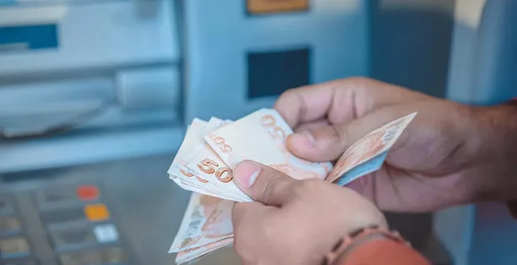 Bir insan iki eliyle 50 TL'lik banknotları sayıyor. Arka planda gri tonlarda para çekme makineleri (ATM) bulunuyor.