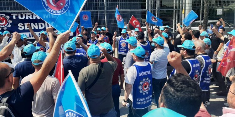 Türk Metal üyesi çok sayıda işçi fabrika önünde toplanmış slogan atıyor. İşçilerin üzerinde Türk Metal önlükleri ellerinde ise sendika bayrakları var.