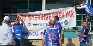 Üzerlerinde Türk Metal sendikasına ait önlükler olan işçiler, fabrikanın demirlerine "Bu işyerinde grev var" yazılı pankart asıyor