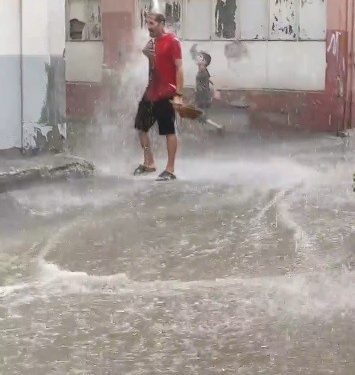 Siyah şortlu, kırmızı tişörtlü bir vatandaş bir binanın oluğundan yola akan yağmur suyunda yıkanıyor.