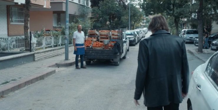 Siyah giyinimli biri arkası dönük hemen önünde arabasıyla domates satan seyyar satıcı bulunuyor.