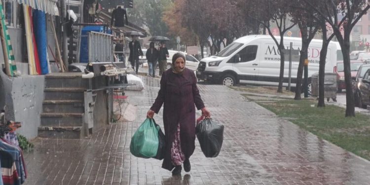 Bir kadın yağmur altında yürüyor. İki elinde de poşet torba bulunmakta.