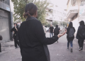Siyah giyinimli bir kadın sokak ortasında durmuş elinde bir taş var. Kadının önünde de eyleme katılan başka kadınlar bulunuyor.