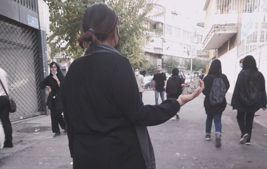 Siyah giyinimli bir kadın sokak ortasında durmuş elinde bir taş var. Kadının önünde de eyleme katılan başka kadınlar bulunuyor.