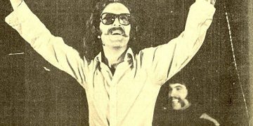 Cem Karaca'nın gençlik döneminde sahnede çekilen bir fotoğrafı. Gözlükleri ile sahne alan ve yüzündeki tebessüm belli olan Karaca, kollarını iki yana açarak şarkı söylüyor.